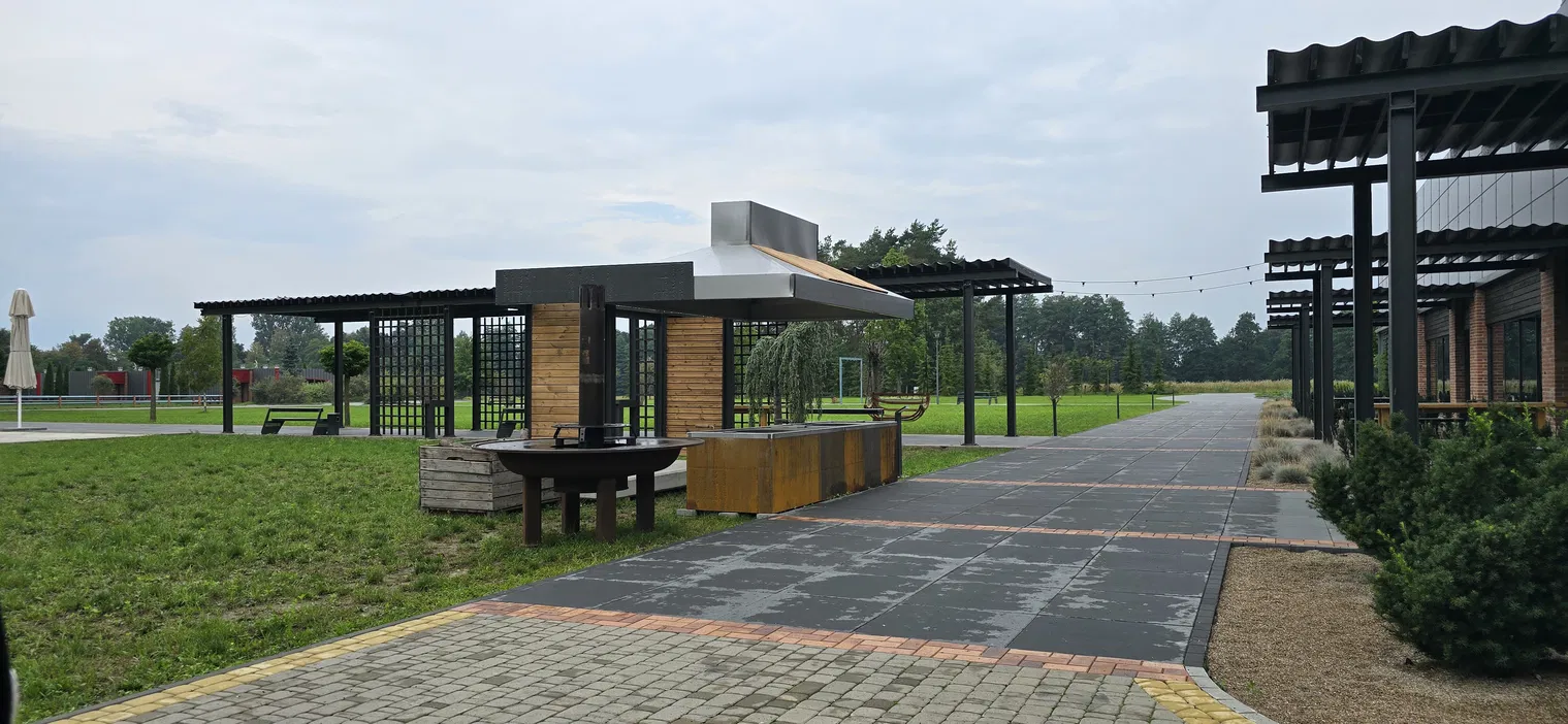 Nowoczesny pawilon ogrodowy z grillem w parku, otoczony zielonym trawnikiem i alejką, w słoneczny dzień.