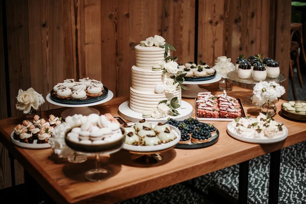 Dekoracyjny stół weselny z eleganckim tortem i różnorodnymi deserami, w tym makaronikami, babeczkami i tartinkami, w stylowej aranżacji na drewnianym tle.