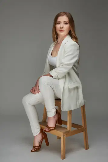  Kobieta w białym garniturze siedząca na krześle, elegancka stylizacja na szarym tle. 