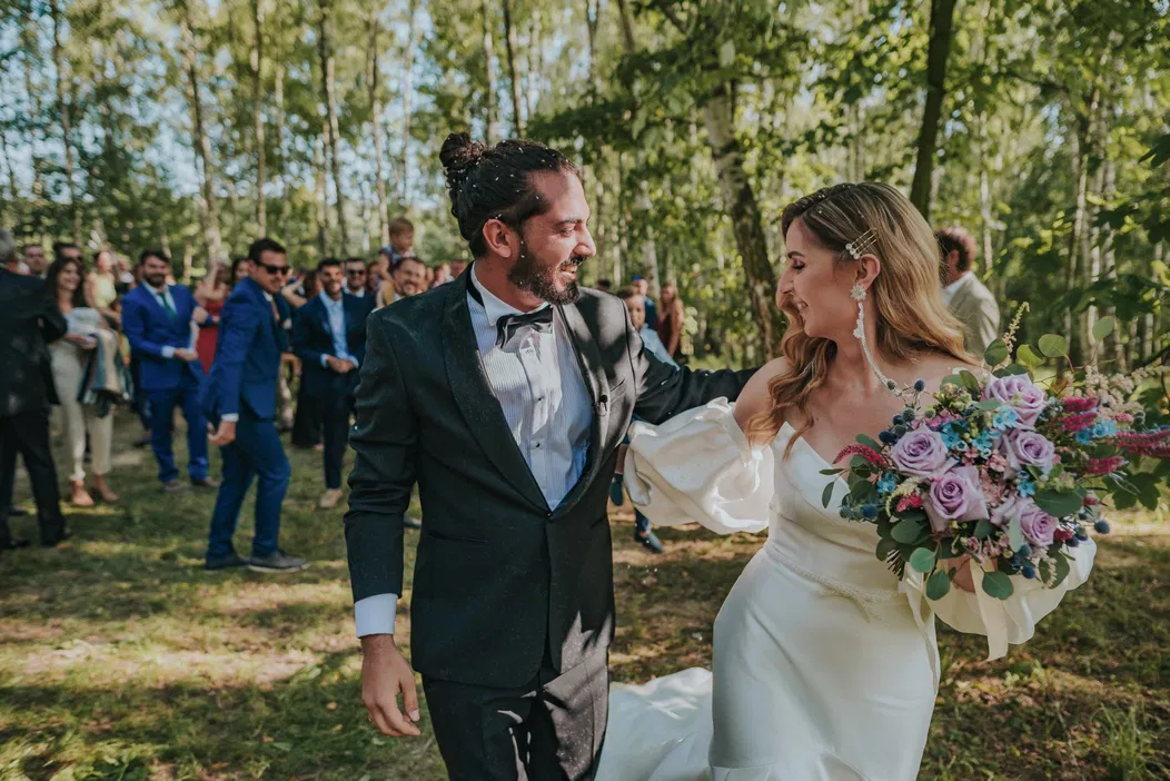 Para młoda podczas wesela w plenerze, uśmiechnięta panna młoda w białej sukni z bukietem kwiatów, pan młody w eleganckim garniturze, romantyczna sceneria wśród drzew, ślub w stylu rustykalnym.