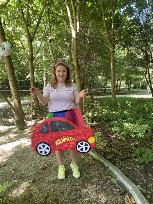 Kobieta w parku z piniatą Hot Wheels w kształcie czerwonego samochodu, drzewa w tle, urodziny Leone’a Stasia.