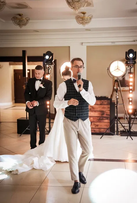 Wodzirej prowadzący wesele w eleganckim wnętrzu z mikrofonem w ręku, z parą młodą w tle.