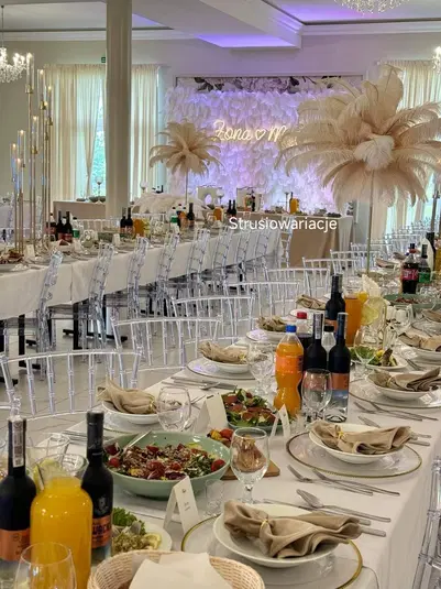  Luksusowa sala weselna z elegancko udekorowanym stołem, ozdoby z piór strusich, białe obrusy, przezroczyste krzesła, świece, napis Żona ❤️ Mąż, ekskluzywne menu, napoje i nastrojowe oświetlenie. 