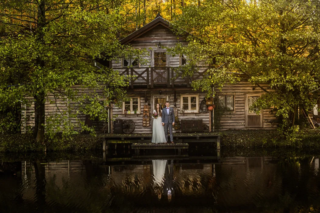 Dwoje młodych ludzi w eleganckich strojach weselnych przed drewnianym domkiem otoczonym drzewami odbijającymi się w spokojnej wodzie.