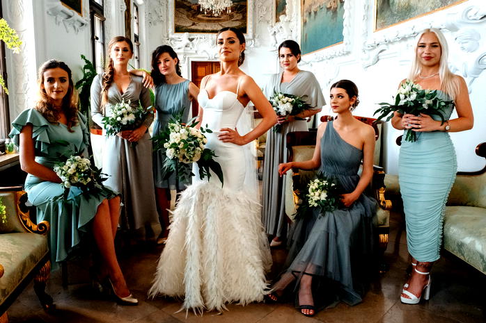  Panna młoda w białej sukni z druhnami w eleganckich sukniach, trzymające bukiety kwiatów, w luksusowym wnętrzu z zabytkowymi detalami. 