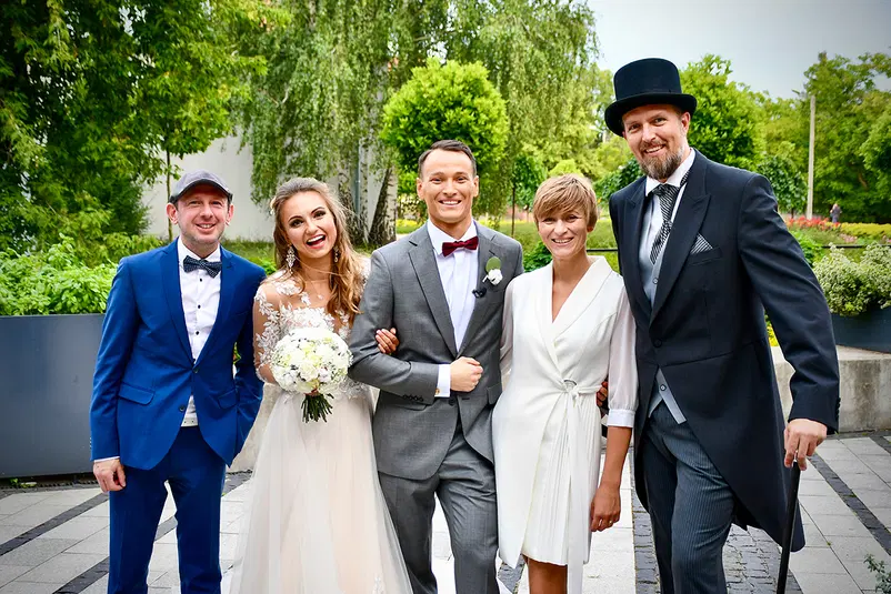  Grupa uśmiechniętych ludzi w eleganckich strojach na świeżym powietrzu, wesele na tle zieleni. 