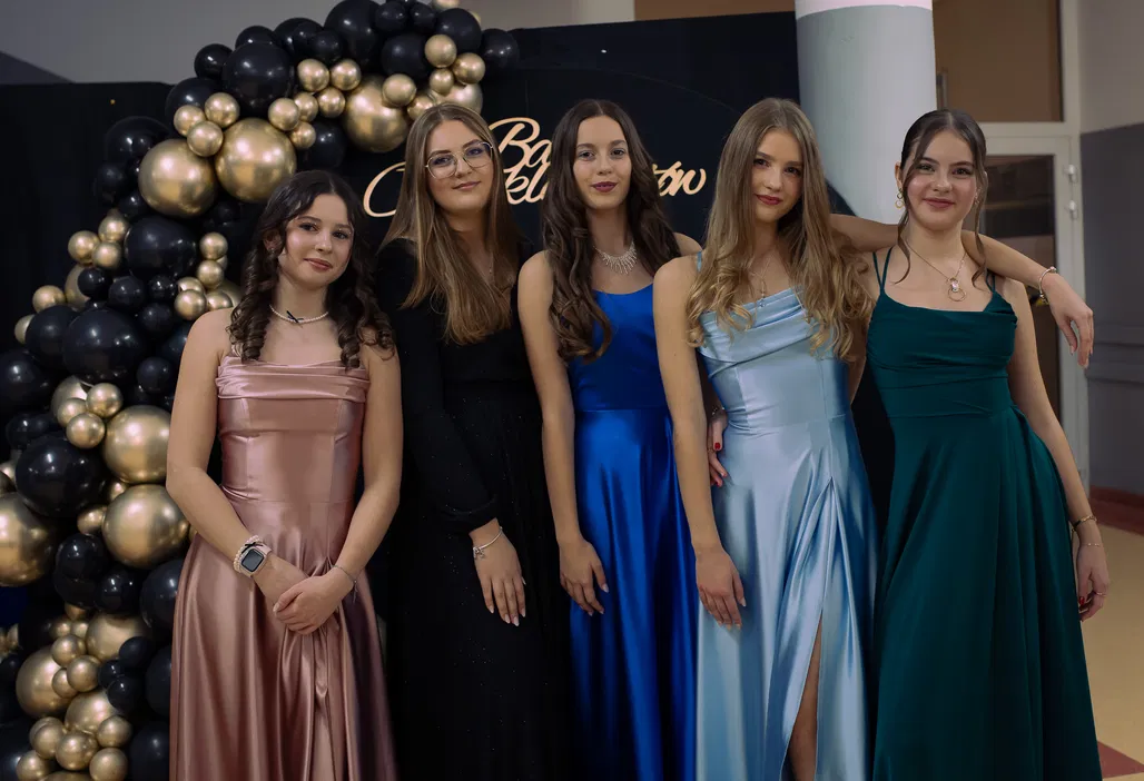 Pięć kobiet w eleganckich sukniach pozujących na tle ozdobnych balonów podczas oficjalnej uroczystości lub balu.
