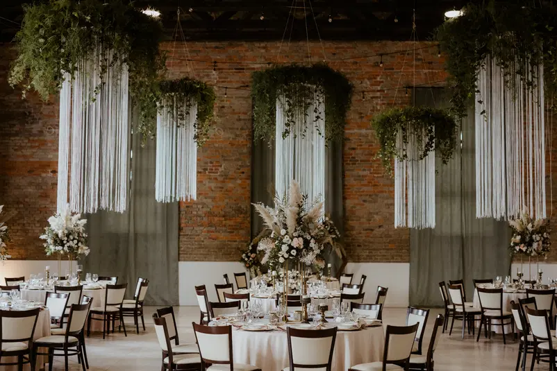  Elegancka sala weselna z okrągłymi stołami, dekoracjami kwiatowymi i stylowymi żyrandolami, na tle ceglanej ściany. 