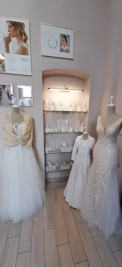  Trzy manekiny z sukniami ślubnymi w eleganckim salonie ślubnym, ozdobione biżuterią i akcesoriami; w tle półka z dodatkami. 
