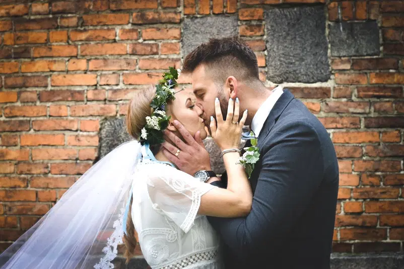  Para młoda całuje się na tle ceglastej ściany, panna młoda w białej sukni i welonie z wiankiem z kwiatów, pan młody w eleganckim garniturze. 