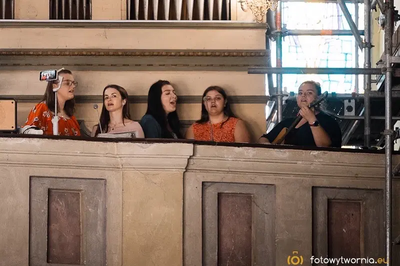  Grupa kobiet śpiewających w kościele, jedna gra na gitarze, wnętrze z witrażami i oświetleniem. 