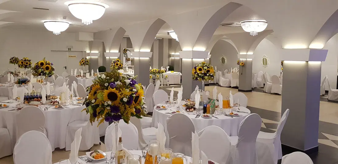  Sala weselna z okrągłymi stołami, eleganckimi białymi obrusami i kwiatowymi aranżacjami z słoneczników oraz innych kwiatów. 