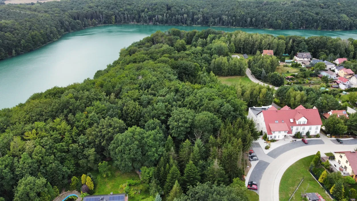 Widok z lotu ptaka na zielony las otaczający jezioro, przy którym znajdują się domy mieszkalne z czerwonymi dachami.