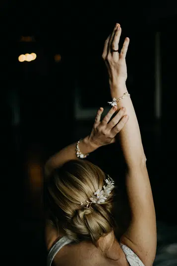  Blondynka w ozdobnej fryzurze z kwiatowymi dodatkami, z podniesionymi rękami w bransoletkach, ciemne tło. 