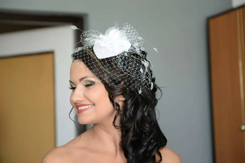  Uśmiechnięta kobieta w eleganckiej fryzurze z ozdobną białą woalką na głowie, patrząca w bok, wnętrze pokoju. 