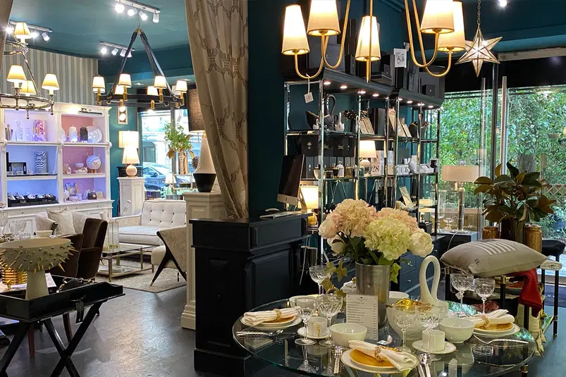  Wnętrze sklepu z eleganckimi meblami i dekoracjami, dobrze oświetlone pomieszczenie z lampami sufitowymi, kwiatami, nowoczesnym stołem udekorowanym porcelaną i szkłem w luksusowym stylu. 