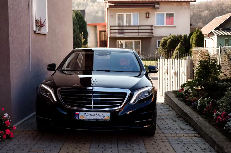  Czarny Mercedes klasy premium z tablicą rejestracyjną Nowożeńcy, zaparkowany na wjeździe przed domem, otoczony kwiatami i zielenią. 