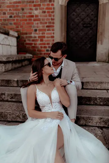  Para młoda w eleganckich strojach ślubnych siedzi na schodach z ceglaną ścianą w tle. 