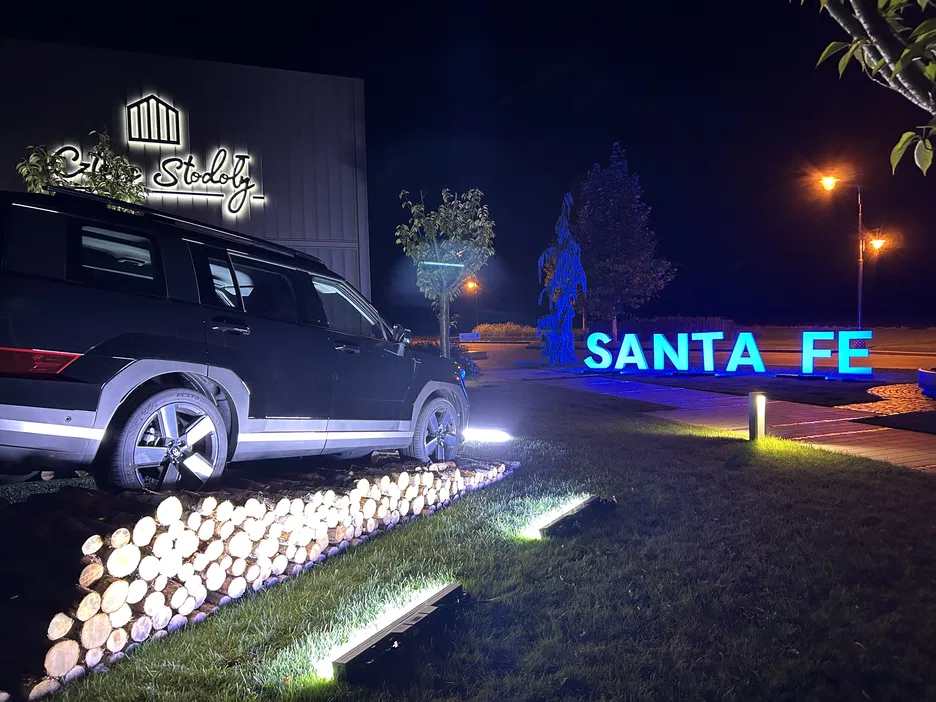 Czarny SUV na tle podświetlonego napisu Santa Fe i budynku Stodoły w nocy, oświetlonego latarniami i reflektorami, na trawniku z ułożonym drewnem.