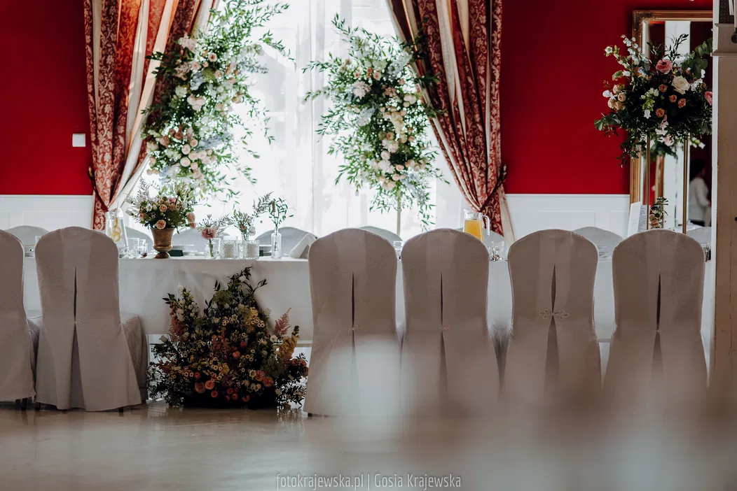 Uroczysta dekoracja weselna z eleganckim bukietem kwiatów na stole, ozdobne krzesła z pokrowcami, stylowe zasłony w czerwieni.