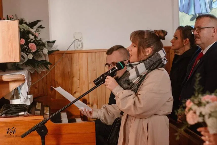 Kobieta śpiewająca w kościele podczas ceremonii, towarzyszy jej mężczyzna grający na organach; w tle osoby uczestniczące w uroczystości.