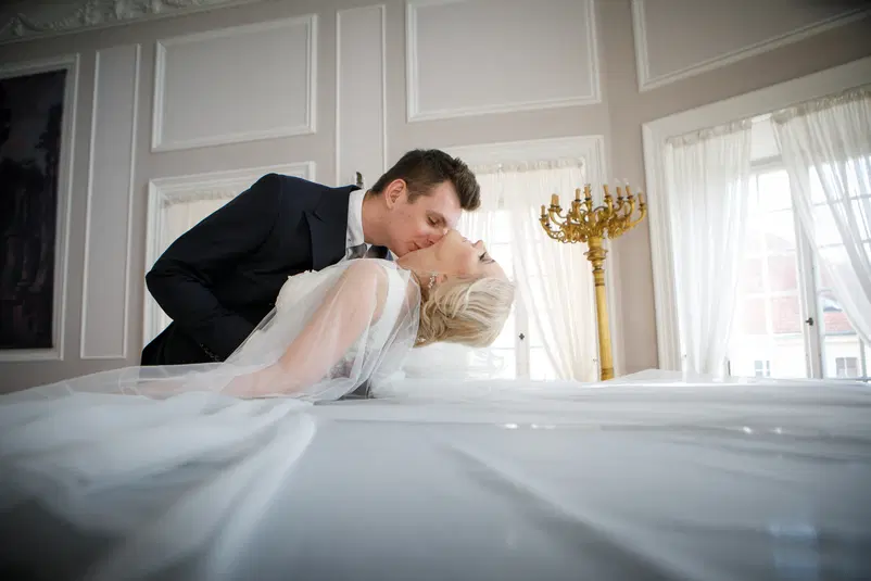  Panna młoda i pan młody całujący się na łóżku w eleganckim wnętrzu z białymi zasłonami i złotym świecznikiem w tle. 