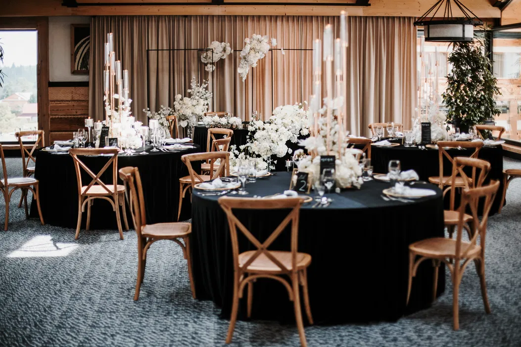 Elegancka sala przyjęć weselnych z okrągłymi stołami, ciemnymi obrusami, drewnianymi krzesłami i kwiatowymi dekoracjami.