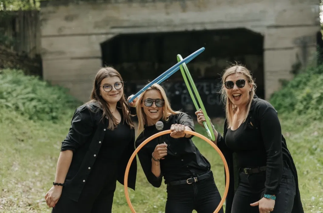 Trzy uśmiechnięte kobiety na świeżym powietrzu, trzymające kolorowe hula hop i balony, noszą okulary przeciwsłoneczne, ubrane na czarno, w tle zielona trawa i betonowa konstrukcja.