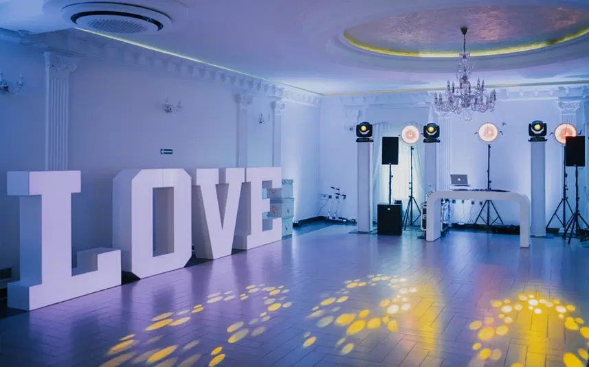  Elegancka sala weselna z dużymi literami LOVE, dekoracją świetlną oraz stanowiskiem DJ-a. 