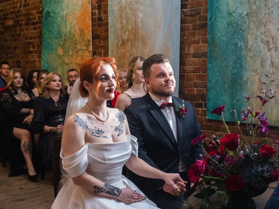 Para młoda podczas ceremonii ślubnej w stylu industrialnym, elegancko ubrani, otoczeni gośćmi w klimatycznej ceglanej sali, dekoracja kwiatowa z czerwonymi różami.