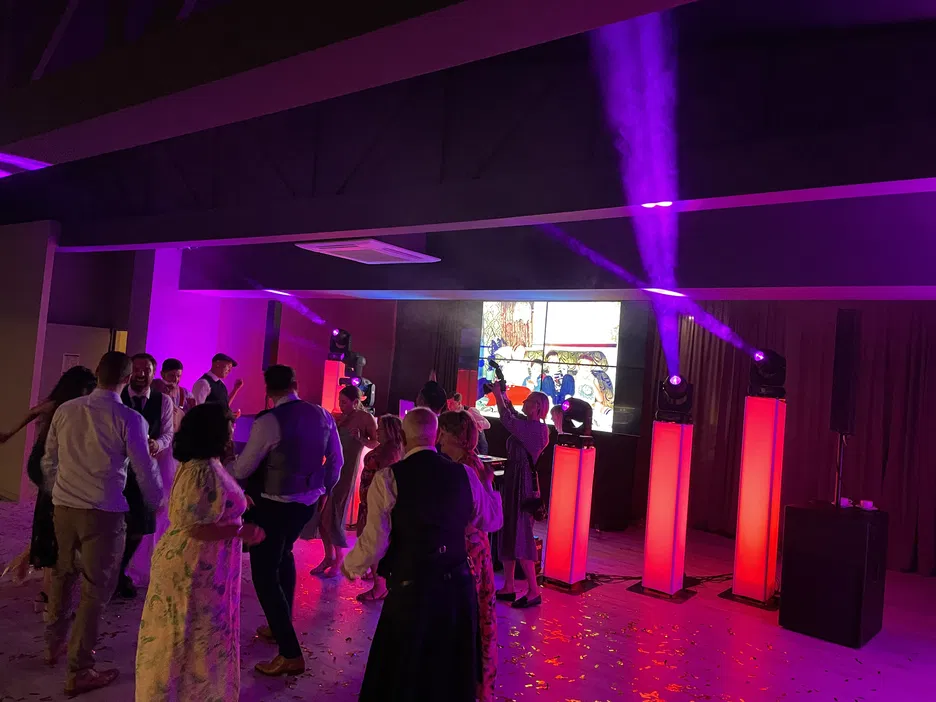 Osoby tańczące na weselu przy kolorowym oświetleniu, scena z DJ-em i ekranem w tle