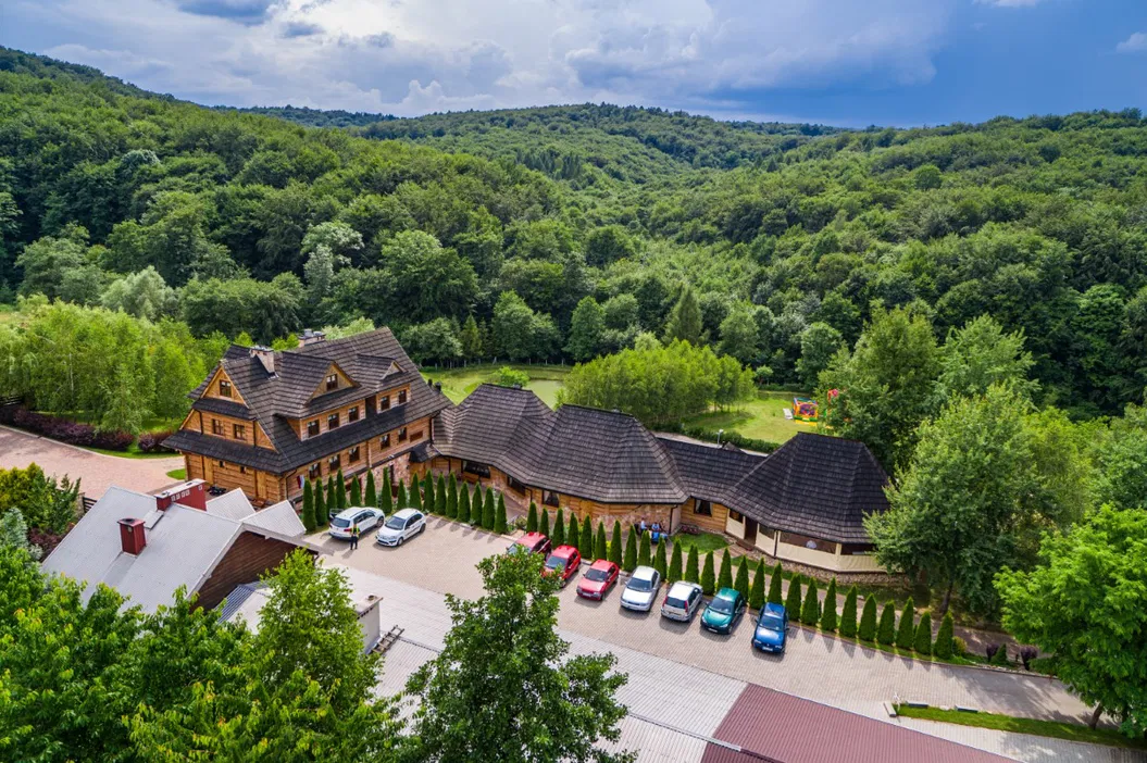Drewniany hotel w górach otoczony lasem, widok z lotu ptaka, malownicza sceneria, parking z samochodami, zielona okolica, turystyka w naturze.