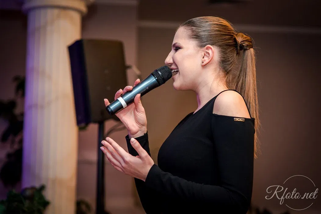 <p>Śpiewająca kobieta na scenie z mikrofonem, koncert muzyczny, elegancka kolumna w tle</p>