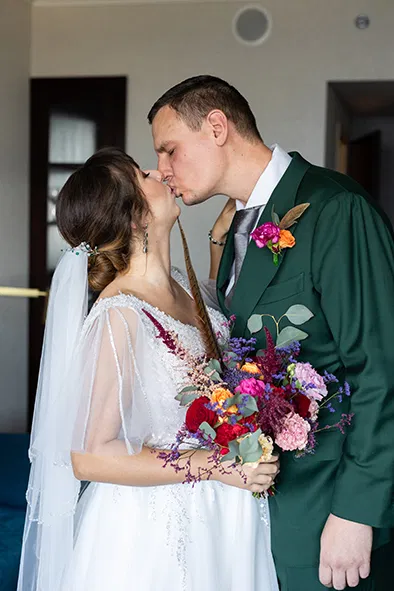 Para młoda całująca się w dniu ślubu, panna młoda w białej sukni z welonem i bukietem kolorowych kwiatów, pan młody w zielonym garniturze.