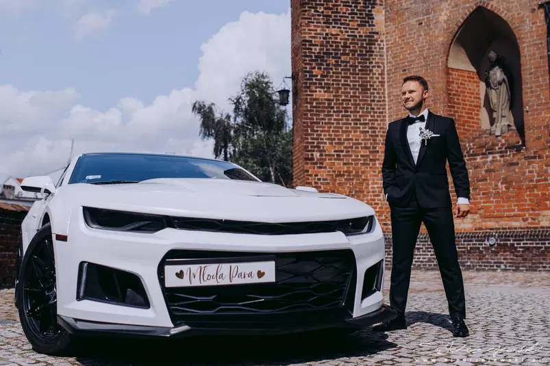  Biały samochód sportowy i elegancko ubrany mężczyzna w garniturze stojący przed zabytkową ceglaną budowlą. 