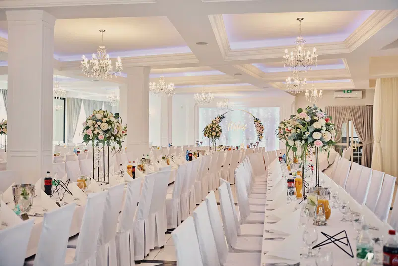  Luksusowa sala weselna z eleganckimi nakryciami stołów, wysokimi bukietami kwiatów i stylowymi żyrandolami. 