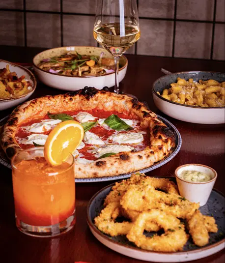  Włoska kolacja: pizza Margherita, kalmary z sosem, makaron i włoskie wino na drewnianym stole, napój pomarańczowy. 