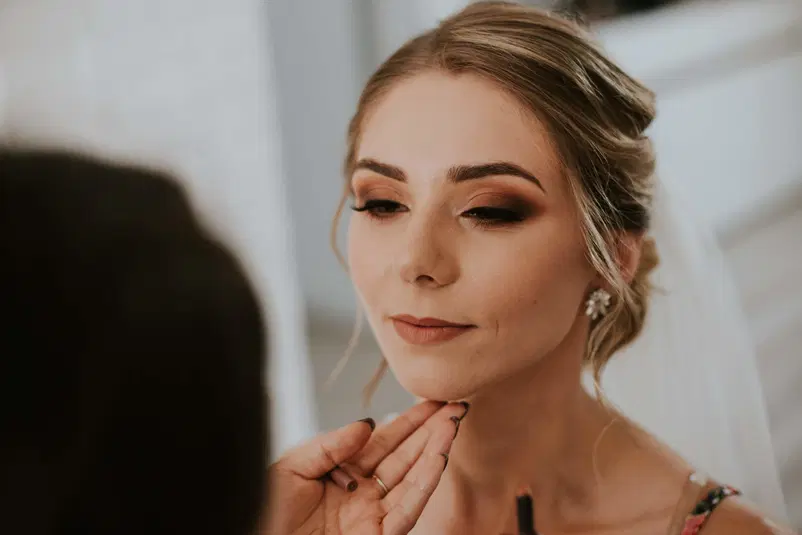  Kobieta w makijażu ślubnym, przygotowywana do ceremonii; profesjonalny makijaż z podkreślonymi oczami i subtelną szminką; stylistka poprawiająca wygląd twarzy; elegancki i klasyczny makijaż ślubny. 