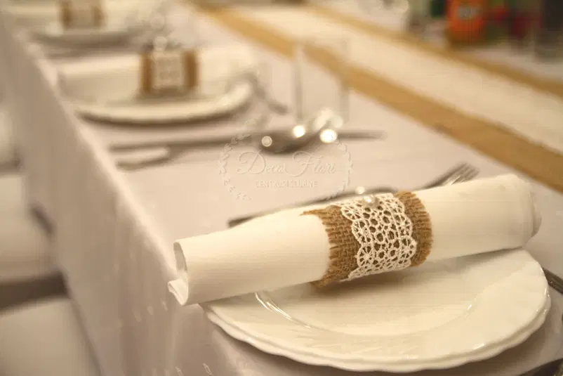  Elegancko przygotowany stół weselny z białymi serwetkami ozdobionymi koronką i jutą na talerzach, minimalistyczna dekoracja ślubna. 
