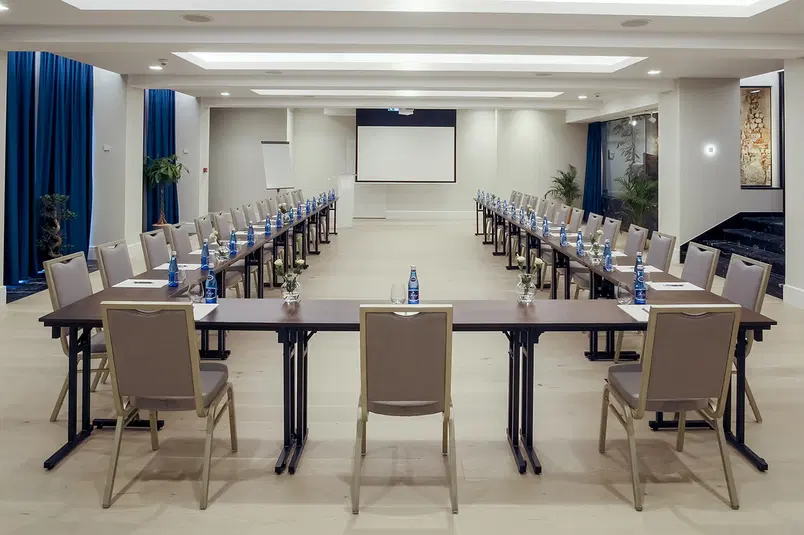  Elegancka sala konferencyjna z ustawieniem w kształcie litery U, wyposażona w krzesła, stoły z wodą, notatnikami i projektorem, idealna na spotkania biznesowe i seminaria. 