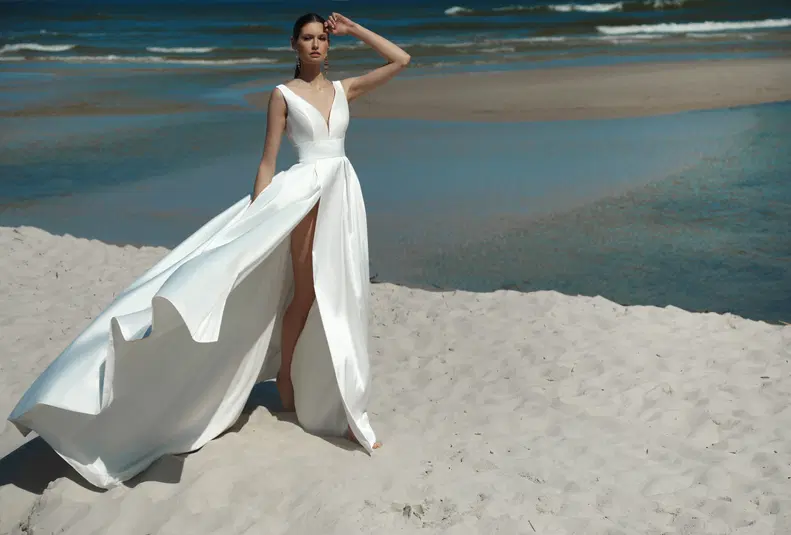  Piękna kobieta w eleganckiej białej sukni z głębokim rozcięciem pozuje na piaszczystej plaży, z falującym morzem w tle. Idealna suknia ślubna na plażowy ślub. 