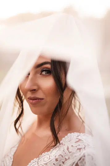  Brunetka w koronkowej sukni ślubnej z woalem, patrząca w kamerę, romantyczny portret panny młodej. 