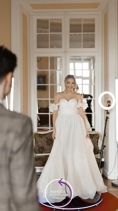 Panna młoda w białej sukni ślubnej, uśmiechająca się w eleganckim wnętrzu z lustrami i sofą, wykorzystanie fotobudki 360.