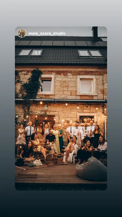 Grupa ludzi podczas przyjęcia na świeżym powietrzu, siedzących i stojących na drewnianym tarasie przed kamiennym budynkiem, z ozdobnymi lampkami oraz zimnymi ogniami, w atmosferze wieczornego spotkania.