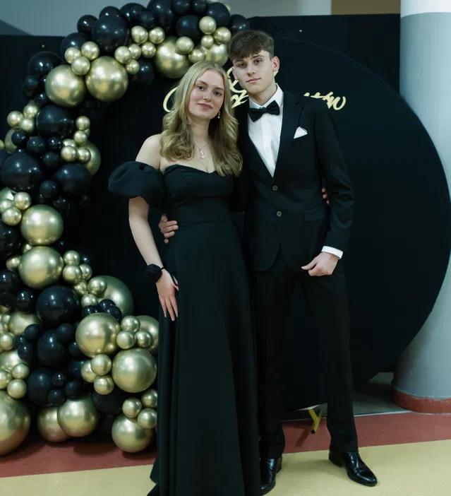 Para w eleganckich strojach na balu, kobieta w czarnej sukni, mężczyzna w garniturze z muchą, tło z czarno-złotymi balonami.