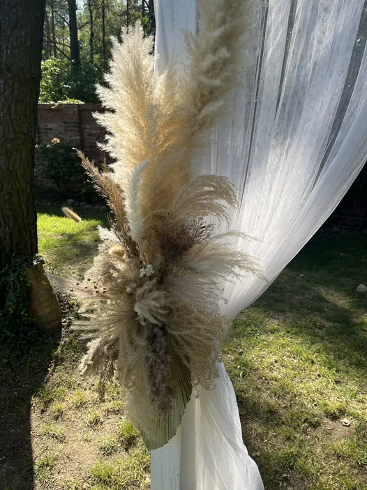 Dekoracja ślubna boho z trawą pampasową na tle białych zasłon w plenerze.