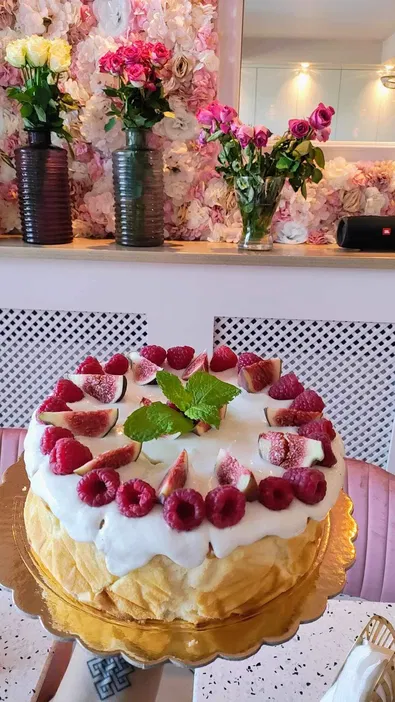 Tort z owocami na złotej podstawce, ozdobiony figami, malinami i miętą, w tle dekoracja kwiatowa i wazon z różami.