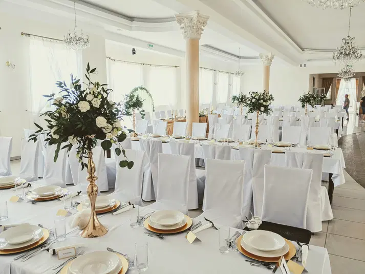  Sala weselna udekorowana na przyjęcie, eleganckie białe pokrowce na krzesła i stoły, wysokie złote wazony z białymi kwiatami i zielenią, zastawiona porcelana oraz sztućce. 