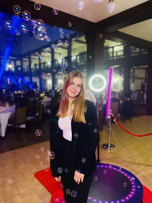 Młoda kobieta w eleganckim stroju w otoczeniu baniek mydlanych na przyjęciu, stojąca na czerwonym dywanie w jasnym i stylowym wnętrzu z neonowymi światłami w tle.