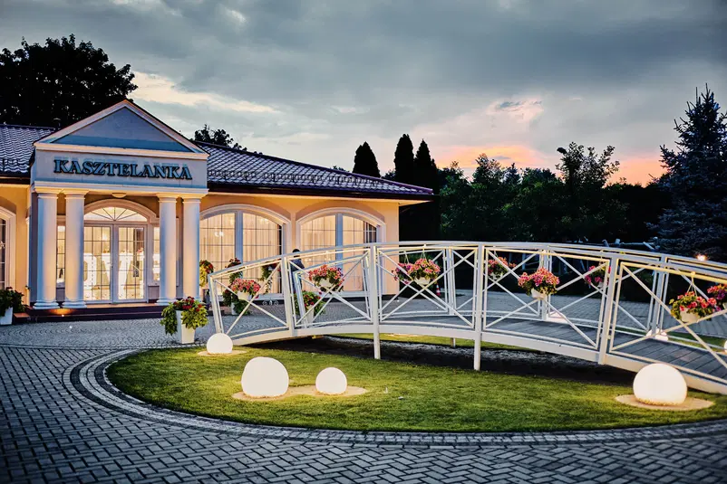  Budynek Kasztelanka z oświetlonym ogrodem i mostkiem, wieczorna sceneria, elegancka architektura. 